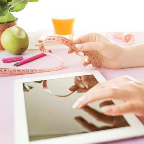 Woman, healthy eating. She working on computer in office. Female hands holding tape. Side view on woman. Trendy color pink desk. Woman, stilish workplace. Tablet, apple. Breakfast. Diet, health concept